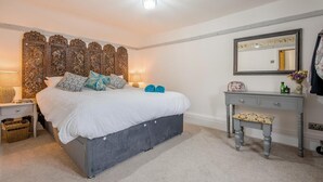 A bedroom with a large bed, a wooden headboard, a bedside table with a lamp, a dresser with a mirror, and a bench. - 2 Bed in Peasmarsh (oc-s33528) (Peasmarsh)