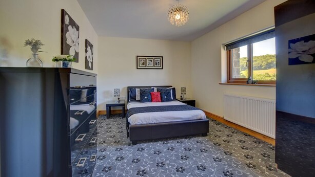 A bedroom with a bed, bedside tables, a dresser, a chandelier, framed pictures, a window with a view, and patterned carpet. - 3 Bed in Aberdyfi (oc-54035) (Pennal)