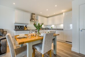 Une cuisine moderne avec des meubles blancs, une table à manger en bois et des chaises recouvertes d’un tissu gris.