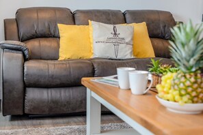 Un canapé en cuir marron agrémenté de deux coussins jaunes et d’un coussin blanc orné d’un logo. Une table basse en bois, sur laquelle reposent deux tasses blanches et un ananas, se trouve devant le canapé.