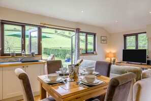A dining area with a table set for four, a television, and a view of a green landscape through large windows.