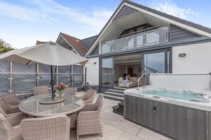 A modern outdoor patio with a glass table, wicker chairs, a hot tub, and a covered seating area. - 4 Bed in Woolacombe (oc-l33535) (Woolacombe)