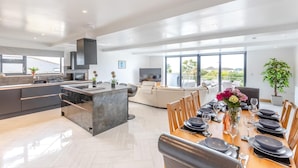 Una cocina moderna con una isla central, comedor con una mesa dispuesta para seis personas y zona de estar con sofá y televisor.