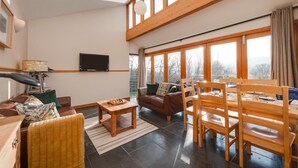 Ein geräumiges Wohnzimmer mit hoher Decke, großen Fenstern, einem Flachbildfernseher, einem Couchtisch aus Holz, einer Couch und einem Essbereich mit Holzsesseln und einem Tisch.