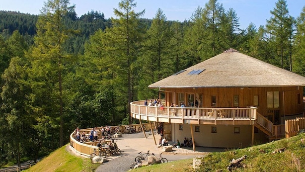 Eine Holzhütte mit einem Balkon und einer runden Sitzecke, umgeben von bewaldeter Landschaft.
