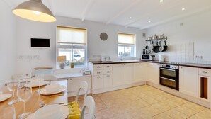 A modern kitchen with white cabinets, a dining table set for four, a large window, and a built-in oven. - 3 Bed in Beadnell (oc-566714) (Swinhoe)