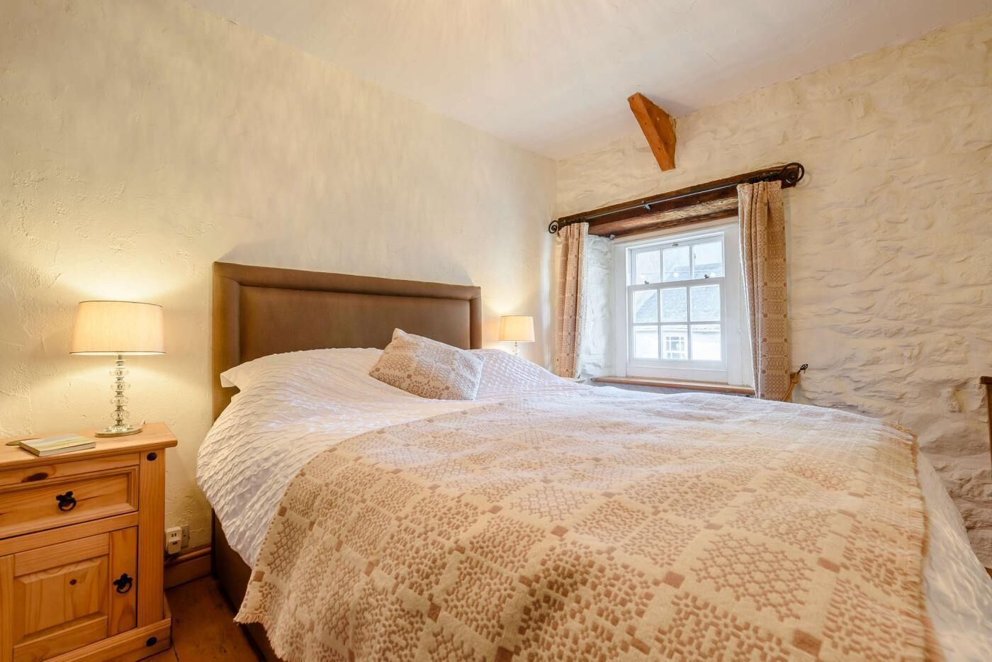 A bedroom with a wooden headboard, a large bed with a patterned blanket, a bedside table with a lamp, and a window with curtains.