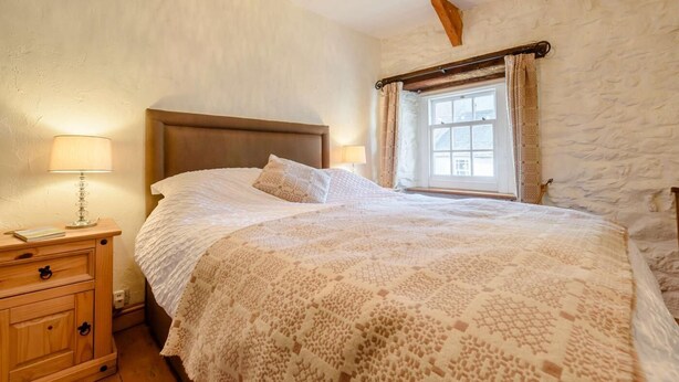 A bedroom with a wooden headboard, a large bed with a patterned blanket, a bedside table with a lamp, and a window with curtains. - 2 Bed in St Davids (oc-30269) (St Davids)