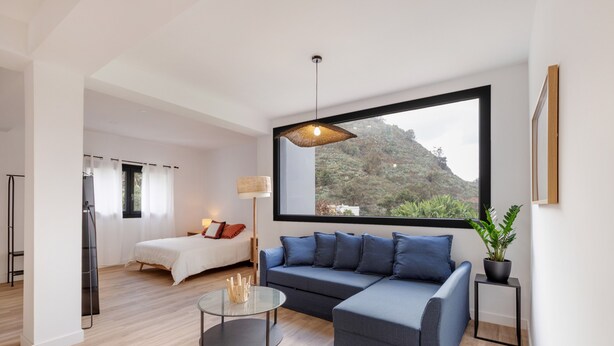 Una sala de estar moderna con sofá, una mesa de centro de vidrio, una cama y un ventanal con vistas a zonas verdes.
