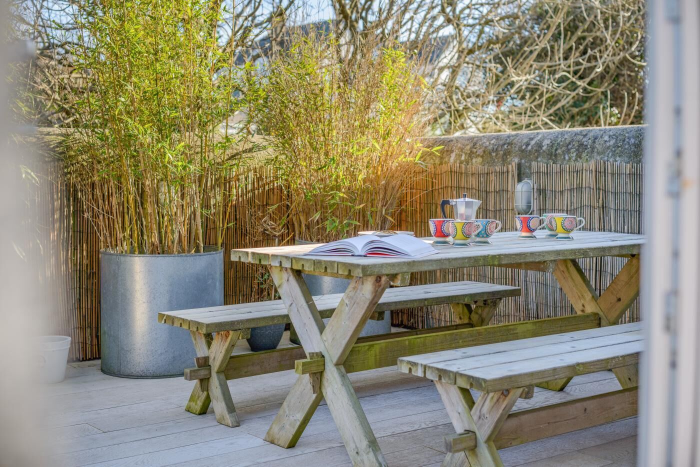 Une table en bois avec des bancs, un livre et des tasses colorées, le tout sur une terrasse agrémentée de plantes en bambou.