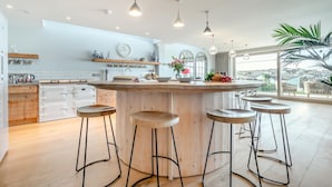 A modern kitchen with a central island, bar stools, and a large window offering a view. - 5 Bed in Polzeath (oc-r33648) (Polzeath)