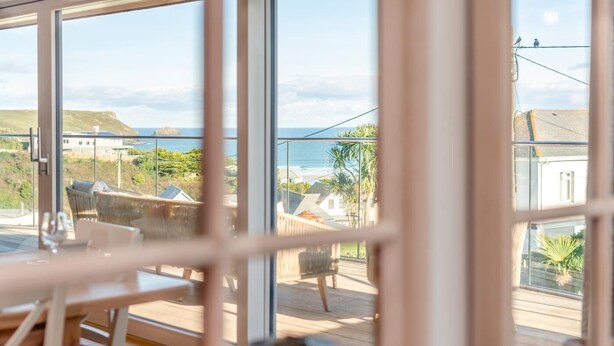 A room with large windows overlooking a deck area with outdoor furniture and a view of the sea. - 5 Bed in Polzeath (oc-r33648) (Polzeath)