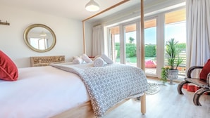 A bedroom with a four-poster bed, a large mirror, a view of the outdoors, and a seating area with a chair and a table. - 5 Bed in Polzeath (oc-r33648) (Polzeath)