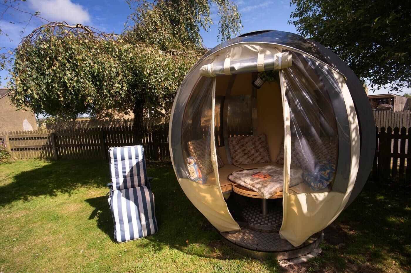 A spherical outdoor tent with a round table and chairs, surrounded by a wooden fence and trees.