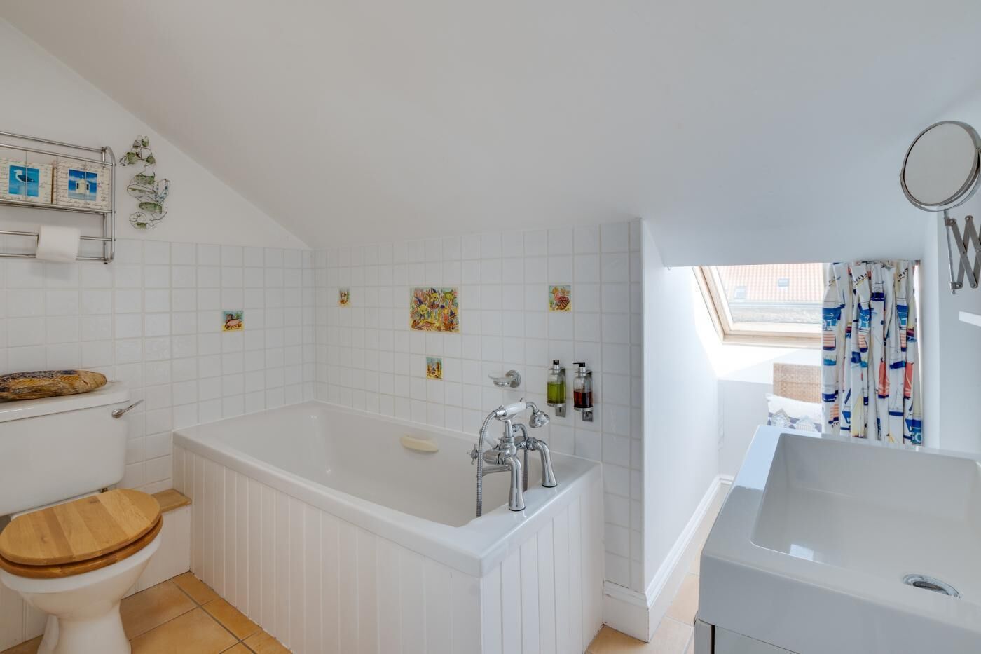 Une salle de bain avec une baignoire blanche, des toilettes avec un abattant en bois et un lavabo. Le mur est décoré de carreaux et il y a une fenêtre avec des rideaux.