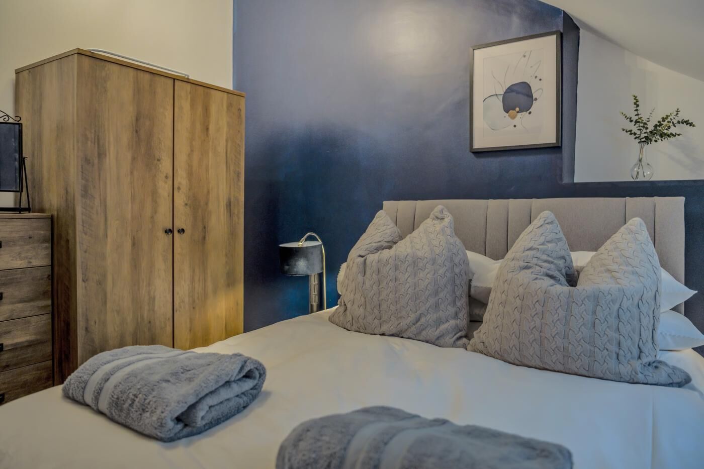 Une chambre à coucher comprenant un lit, des oreillers, une armoire, une œuvre d’art accrochée au mur et un vase avec des plantes.