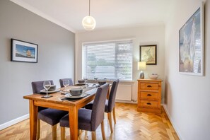 A dining area with a wooden table set for six, flanked by chairs, a window with blinds, a radiator, and a wooden cabinet with drawers.