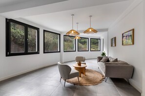 Habitació àmplia amb grans finestres, una taula rodona de fusta, dues cadires, un sofà i lluminàries penjants.