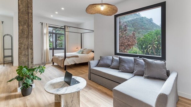A modern room with a bed, a sofa, a large window with a view of greenery, a hanging light fixture, and a laptop on a table. - Vacation Apartment '5' with Mountain View, Shared Terrace and Shared Garden (Moya)