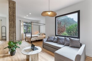 Una habitación moderna con una cama, un sofá, un ventanal con vistas a la vegetación, una lámpara colgante y un ordenador portátil sobre una mesa.