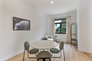 Una habitación con una mesa redonda, cuatro sillas, un sillón individual, una estantería alta de metal negro, un cuadro en la pared y una ventana con cortinas.
