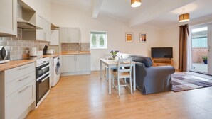 A modern kitchen with white cabinets, a dining area with a table and chairs, a gray sofa, and a TV in a living room. - 3 Bed in Chedgrave (oc-2252) (Chedgrave)