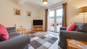 A living room with a sofa, a coffee table, a TV on a stand, a radiator, and a sliding glass door leading to an outdoor area with potted plants. - 3 Bed in Chedgrave (oc-2252) (Chedgrave)