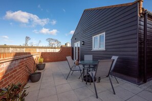 A patio with a table and chairs, a brick wall, and a dark wooden building. - 3 Bed in Chedgrave (oc-2252) (Chedgrave)