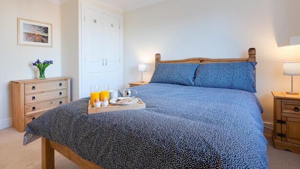 A neatly made bed with a blue patterned bedspread, a bedside table with a lamp, and a wooden dresser with a vase of flowers.