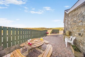 A patio area with a wooden table and chairs, a stone wall, and a view of the countryside. - 3 Bed in Aberystwyth (oc-76692) (Llanilar)