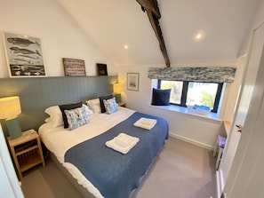 Une chambre à coucher avec un lit, des oreillers, une table de chevet, une fenêtre donnant sur un beau paysage et un plafond avec des poutres apparentes.