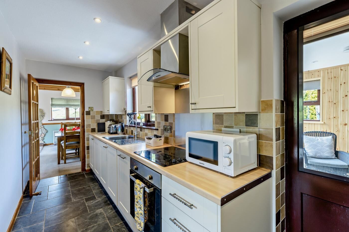 Une cuisine moderne avec des meubles blancs, un four à micro-ondes et une cuisinière. On aperçoit un coin repas avec une table et des chaises à travers une porte ouverte.
