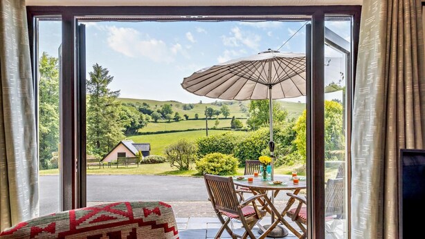 A room with a sliding glass door leading to an outdoor patio. The patio has a table with chairs, a large umbrella, and a view of a rural landscape with fields and a small building. - 2 Bed in Aberystwyth (oc-99869) (Pontarfynach)