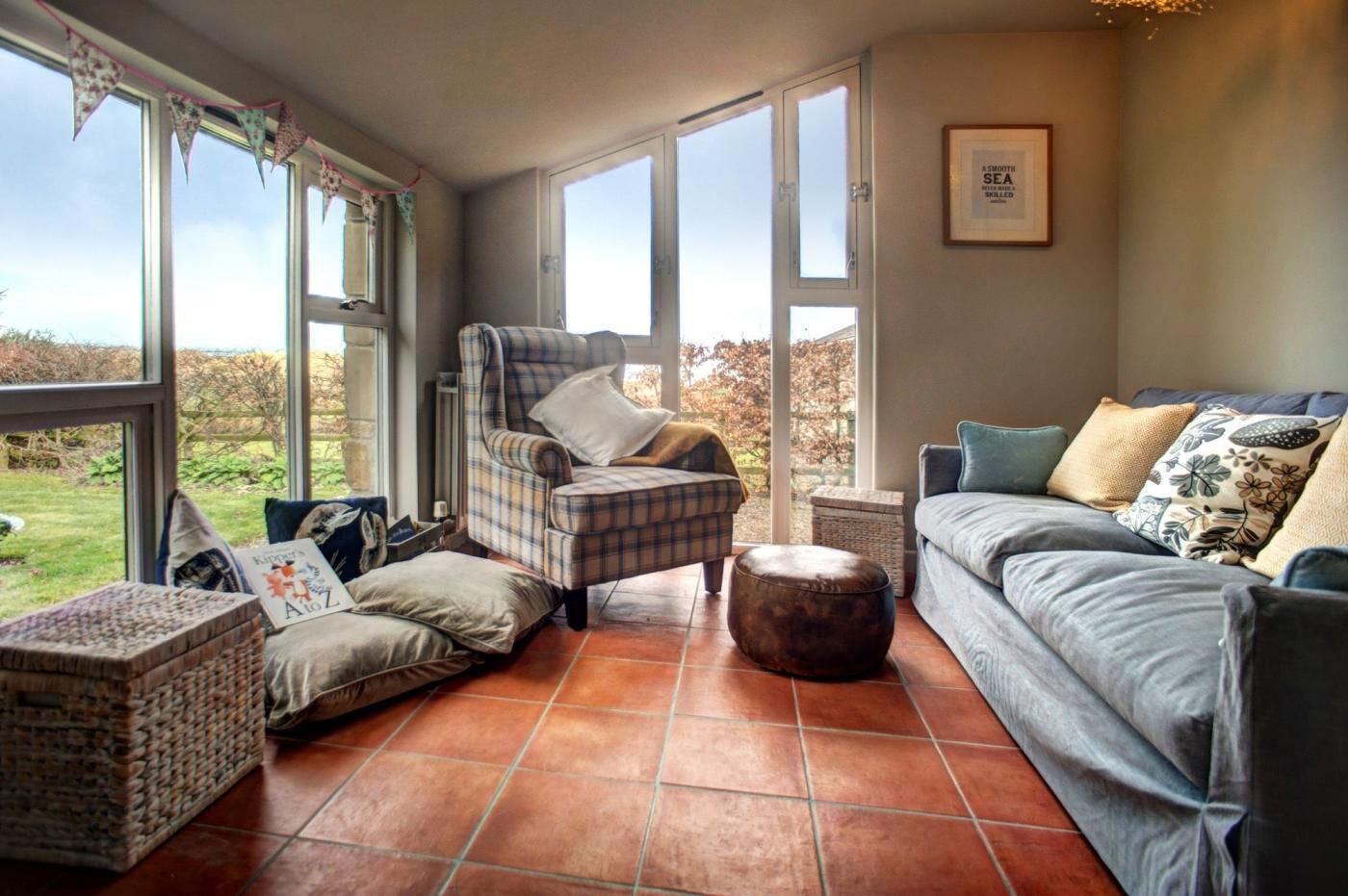 A cozy living room with a plaid armchair, a grey sofa, and a small ottoman. Large windows offer a view of the outdoors.