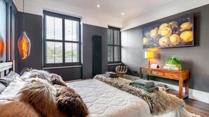 A modern bedroom with a large bed, a wooden nightstand, a painting, and a window with blinds. - 2 Bed in Holmfirth (oc-f33379) (Holmfirth)