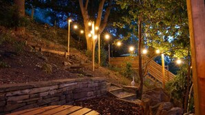 A wooden deck with stone steps leading up a slope, surrounded by trees and illuminated by string lights. - 2 Bed in Holmfirth (oc-f33379) (Holmfirth)