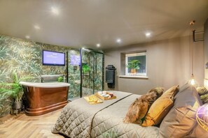 Quarto de hotel moderno com uma cama grande, uma receção de madeira, uma televisão e uma janela com uma planta.