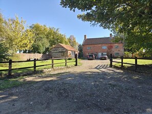A gravel driveway leading to a brick house with a red roof, surrounded by a wooden fence and trees.