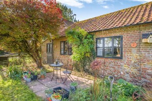 A brick house with a tiled roof, a garden with potted plants, and a small outdoor table and chairs.