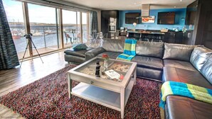 Een moderne woonkamer met een groot raam, een leren bank, een glazen salontafel, en een keuken op de achtergrond.