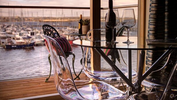 Een glazen tafel met stoelen op een balkon met uitzicht op een jachthaven vol boten.