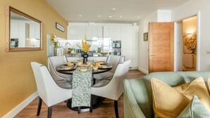 A modern dining area with a round table set for four, surrounded by white chairs. A kitchen with white cabinets and stainless steel appliances is visible in the background. - 2 Bed in Torquay (oc-o33463) (Torquay)