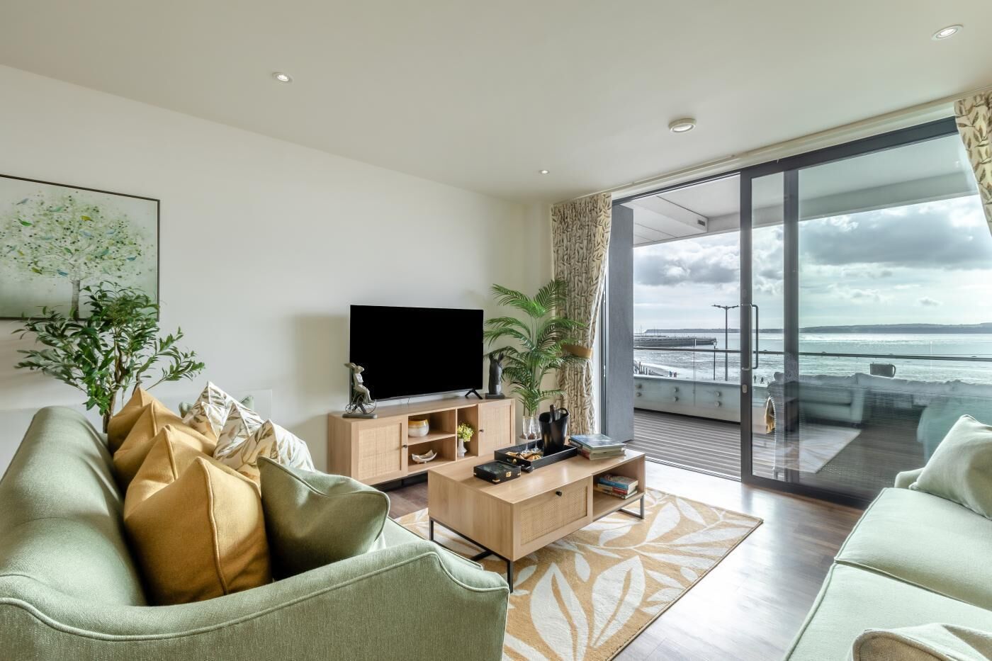 A modern living room with a large TV, wooden furniture, and a view of the sea through large glass doors.