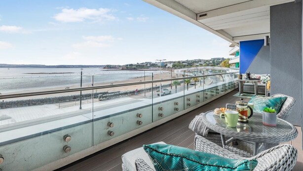 A balcony with a glass railing overlooking a beach, wicker furniture, and a view of the sea and distant buildings. - 2 Bed in Torquay (oc-o33463) (Torquay)