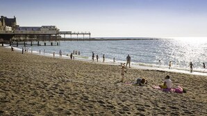 Een strand met mensen die wandelen en zitten op het zand, een pier op de achtergrond en een heldere lucht.