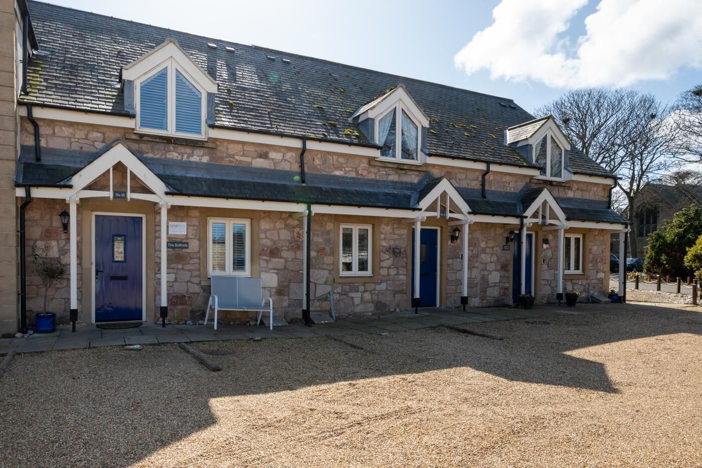 A row of stone buildings with multiple doors and windows, a gravel driveway, and a bench in front.