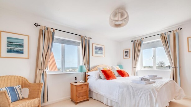 A bedroom with a large bed, two bedside tables, a nightstand, a wicker chair, and a view of the sea through the windows. - 3 Bed in Seahouses (oc-566761) (Seahouses)
