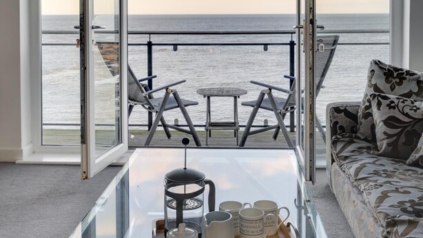 A room with a large glass table, a sofa, and a view of the sea through open windows. - 4 Bed in Beadnell (oc-566397) (Beadnell)