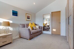 A modern bedroom with a grey sofa, a bedside table with a lamp, a wooden door, and a mirror reflecting the room. - 4 Bed in Beadnell (oc-566397) (Beadnell)