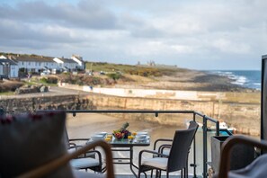 A terrace with a table set for dining, overlooking a beach and houses. - 3 Bed in Craster (oc-566727) (Dunstan)